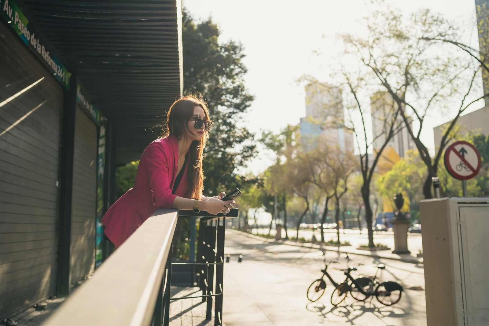 A woman leaning on a railing to check her phone on a sunny street in Mexico City