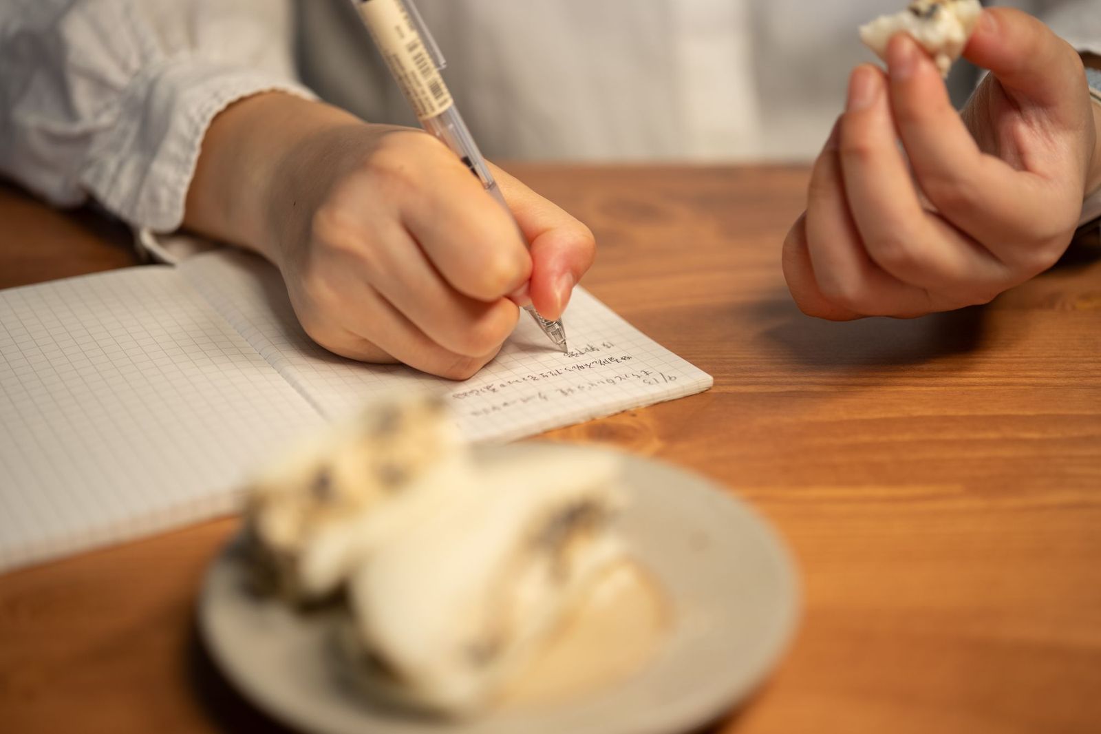 A hand writing notes in a grid notebook on a wooden table, with a snack nearby